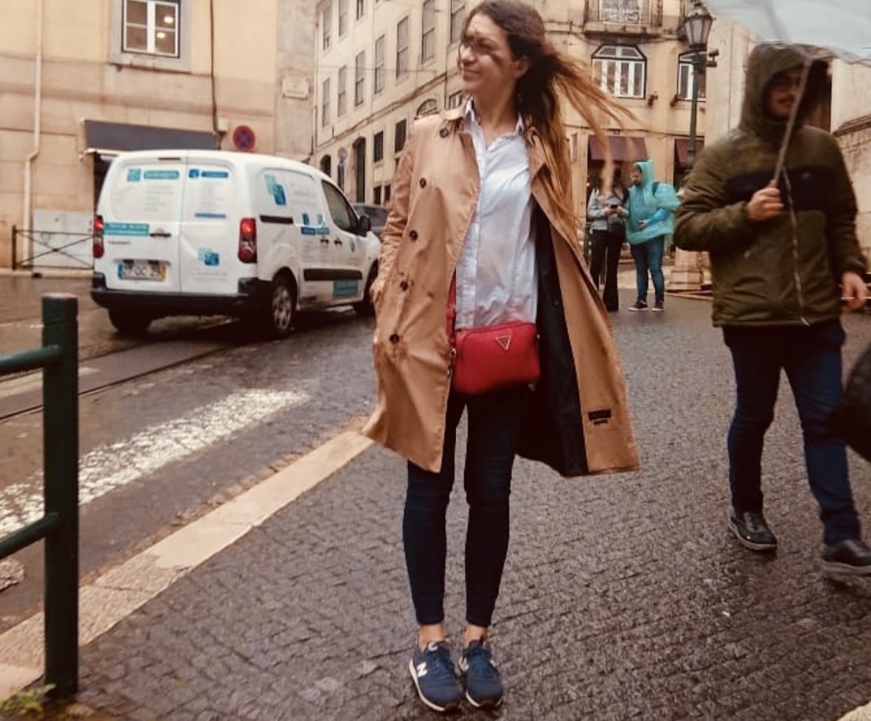 Woman in a trench coat standing on a wet Lisbon street with cobblestones during a rainy day.