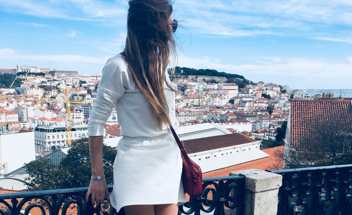 A woman in a white dress looking at a panoramic city view of Lisbon’s red rooftops and historical hills under a blue sky.
