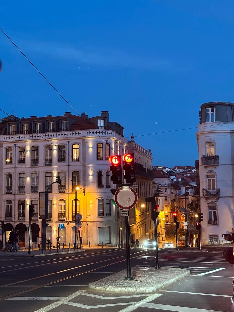 Historic Lisbon corner buildings illuminated at dusk in the Estrela neighborhood, with empty tram tracks and warm streetlights.