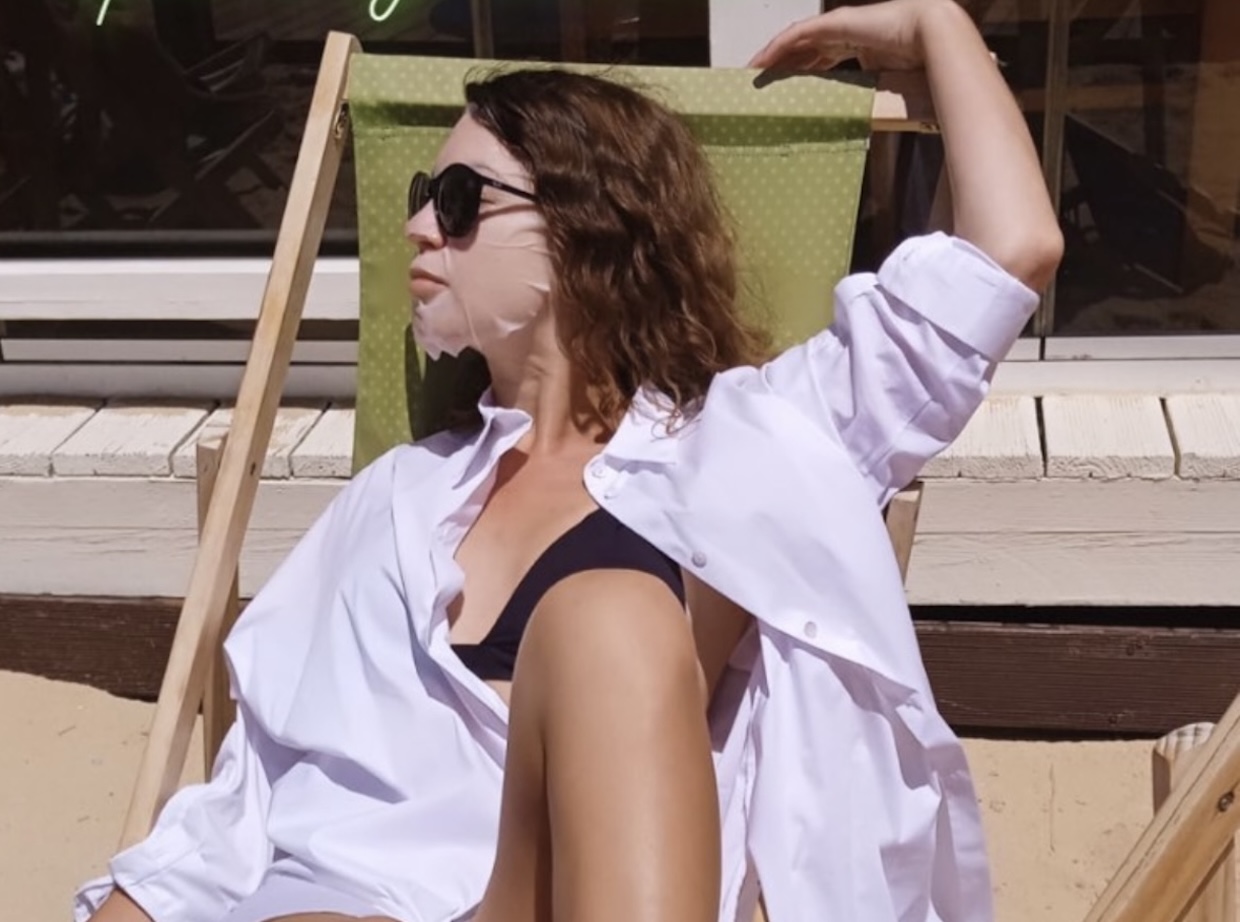 Close-up of a woman in sunglasses and a sheet mask relaxing in a sun lounger in Lisbon, wearing a white oversized shirt.