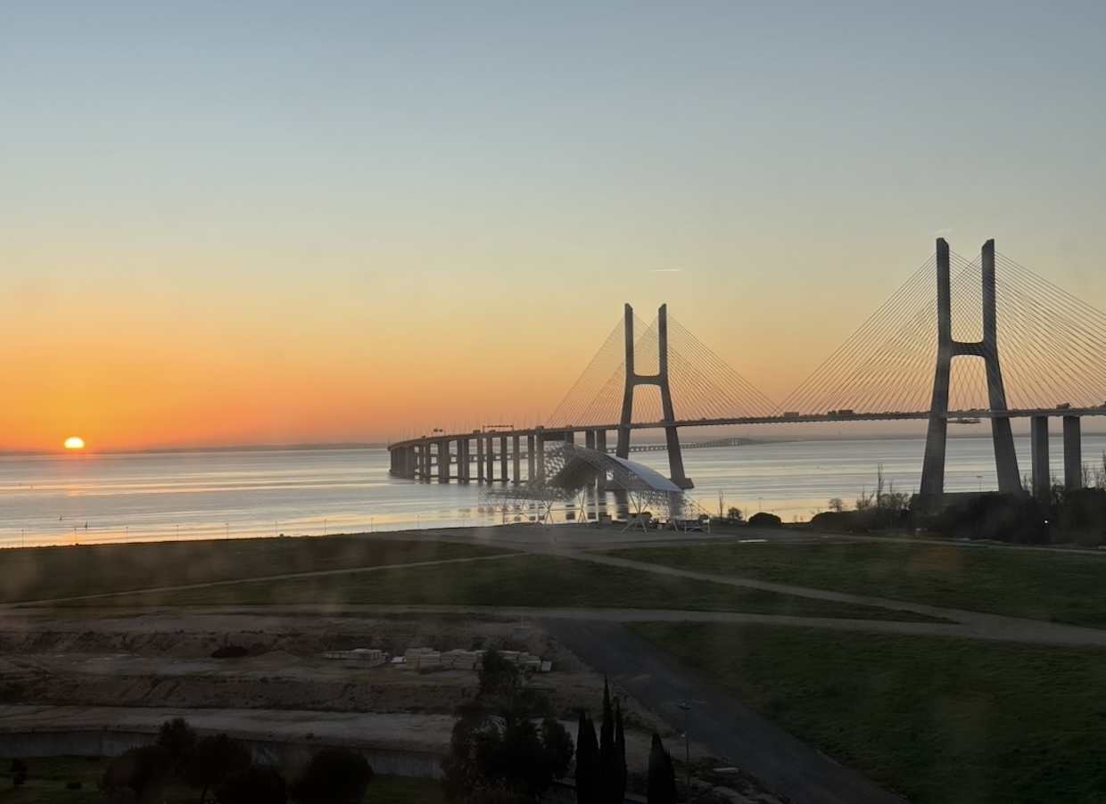 Sunrise over the Vasco da Gama Bridge in Lisbon, with calm water, an orange sky, and the bridge stretching across the horizon.
