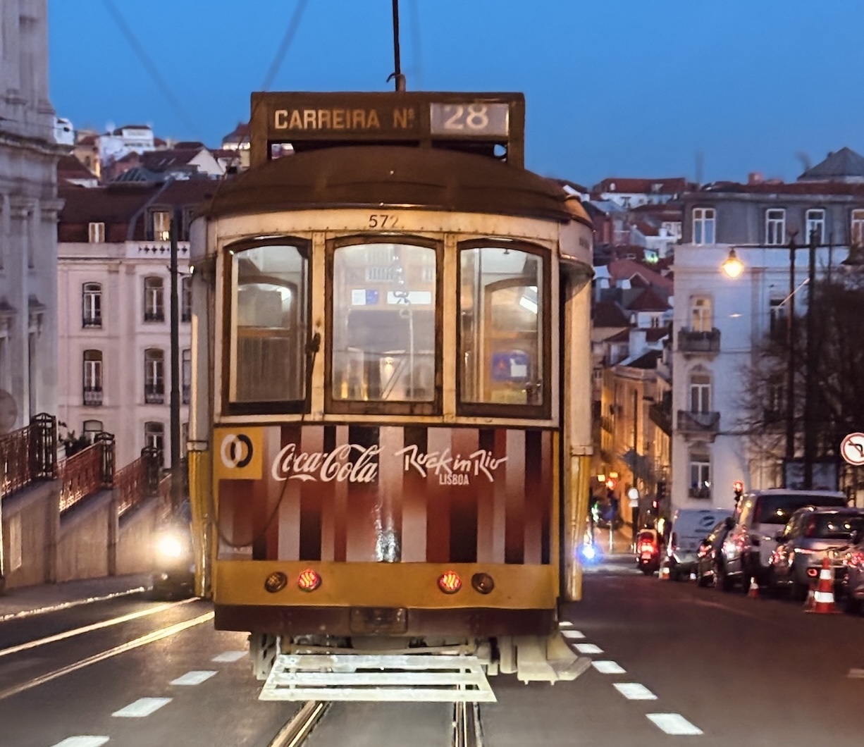 The iconic yellow Tram 28 in Lisbon at night, illuminated by city lights against a deep blue twilight sky, moving through the historical streets.
