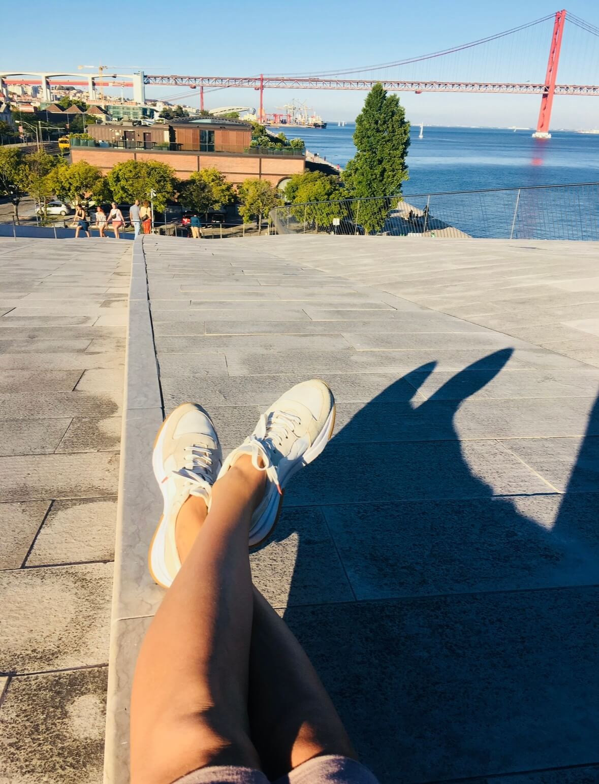 Point-of-view shot of relaxed legs resting on the architectural white roof of the MAAT museum in Belém, Lisbon. The iconic 25 de Abril Bridge and the Tagus River are visible in the background. An inspiring outdoor workspace for creatives and digital nomads.