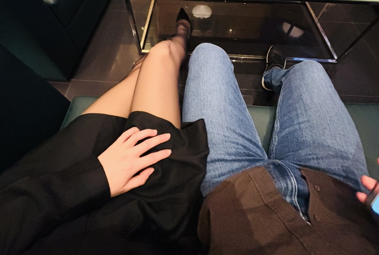 A POV shot of a couple’s legs, with a woman in a black dress, pantyhose, and black heels resting her hand on her partner’s knee during a date in Lisbon.